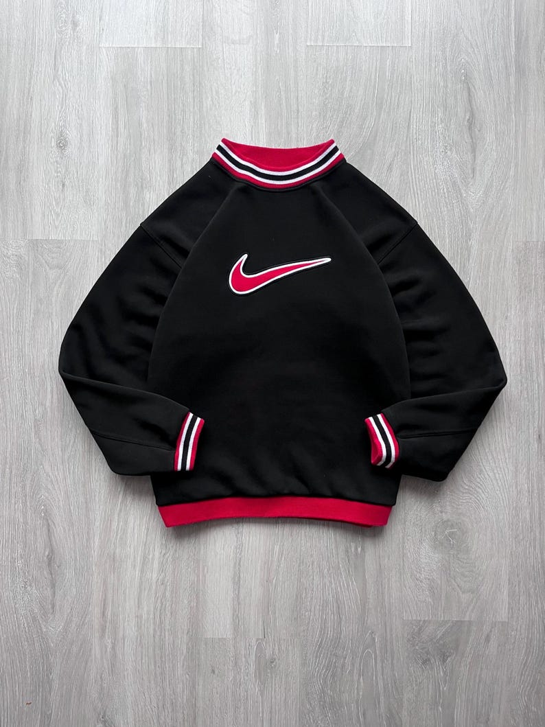May include: Black sweatshirt with red and white striped detailing on the collar, cuffs, and hem. A red Nike swoosh logo, outlined in white, is on the chest. The long-sleeved top has a crew neck.