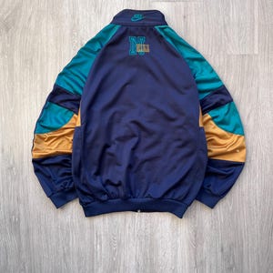 May include: A vintage Nike track jacket in navy blue, teal, and gold. The jacket features a full zipper, a stand-up collar, and the Nike logo on the chest and collar. The back of the jacket has a large "N" and "NIKE" logo.