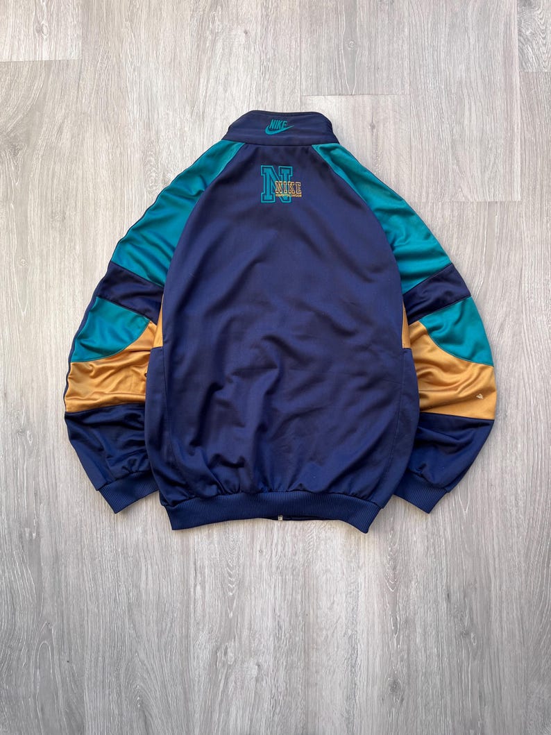 May include: A vintage Nike track jacket in navy blue, teal, and gold. The jacket features a full zipper, a stand-up collar, and the Nike logo on the chest and collar. The back of the jacket has a large "N" and "NIKE" logo.