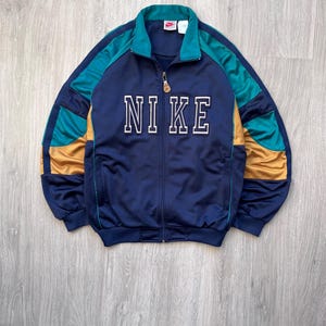 May include: Navy blue track jacket with the word "NIKE" in white block letters across the chest. The jacket features teal and gold accents on the sleeves and collar. It has a full zipper closure and a small Nike logo on the collar.