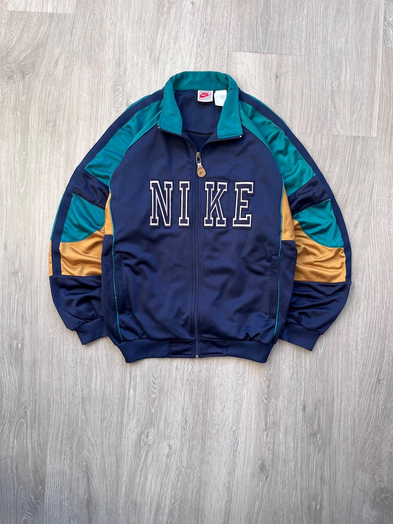May include: Navy blue track jacket with the word "NIKE" in white block letters across the chest. The jacket features teal and gold accents on the sleeves and collar. It has a full zipper closure and a small Nike logo on the collar.