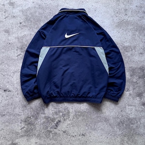 May include: A navy blue windbreaker jacket featuring a white Nike swoosh logo embroidered on the upper back. The jacket includes a collar, long sleeves with elastic cuffs, and light gray side panels accented by a thin white horizontal piping detail.