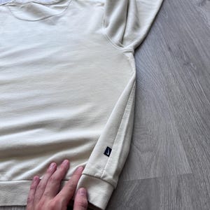 Puede incluir: Sudadera color crema con mangas largas. Tiene cuello redondo y un peque&ntilde;o logotipo negro en el pu&ntilde;o izquierdo. La tela parece suave y c&oacute;moda.