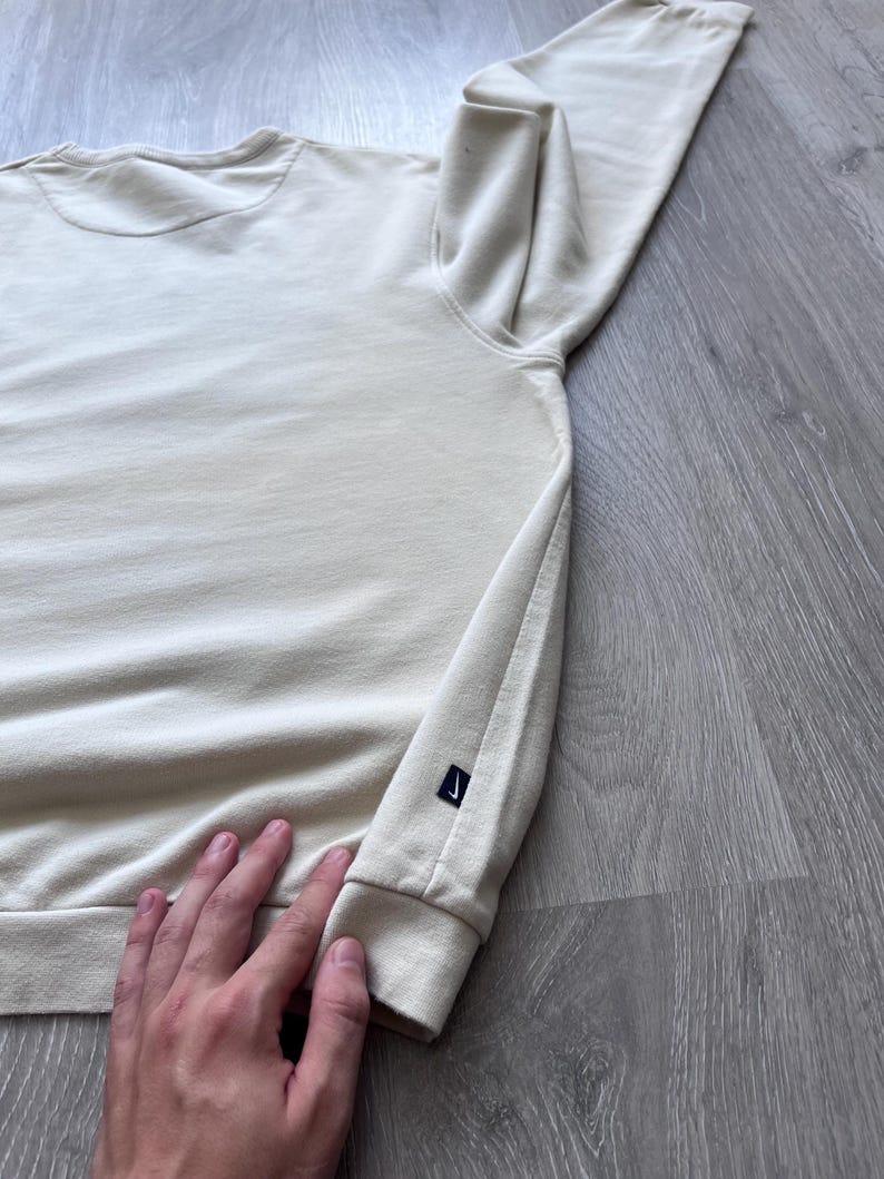 Puede incluir: Sudadera color crema con mangas largas. Tiene cuello redondo y un peque&ntilde;o logotipo negro en el pu&ntilde;o izquierdo. La tela parece suave y c&oacute;moda.