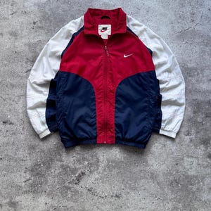 Può includere: Una giacca a vento Nike con zip caratterizzata da un design a blocchi di colore rosso, blu navy e bianco. La giacca presenta un colletto alto, una chiusura frontale con zip integrale e un piccolo logo Nike swoosh bianco sul petto a sinistra.