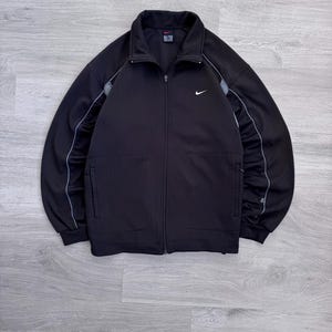 Puede incluir: Chaqueta deportiva negra con cremallera y logotipo blanco de Nike en el pecho. La chaqueta tiene cuello alto, dos bolsillos delanteros y rayas grises en las mangas. Fabricada con un material suave y deportivo.