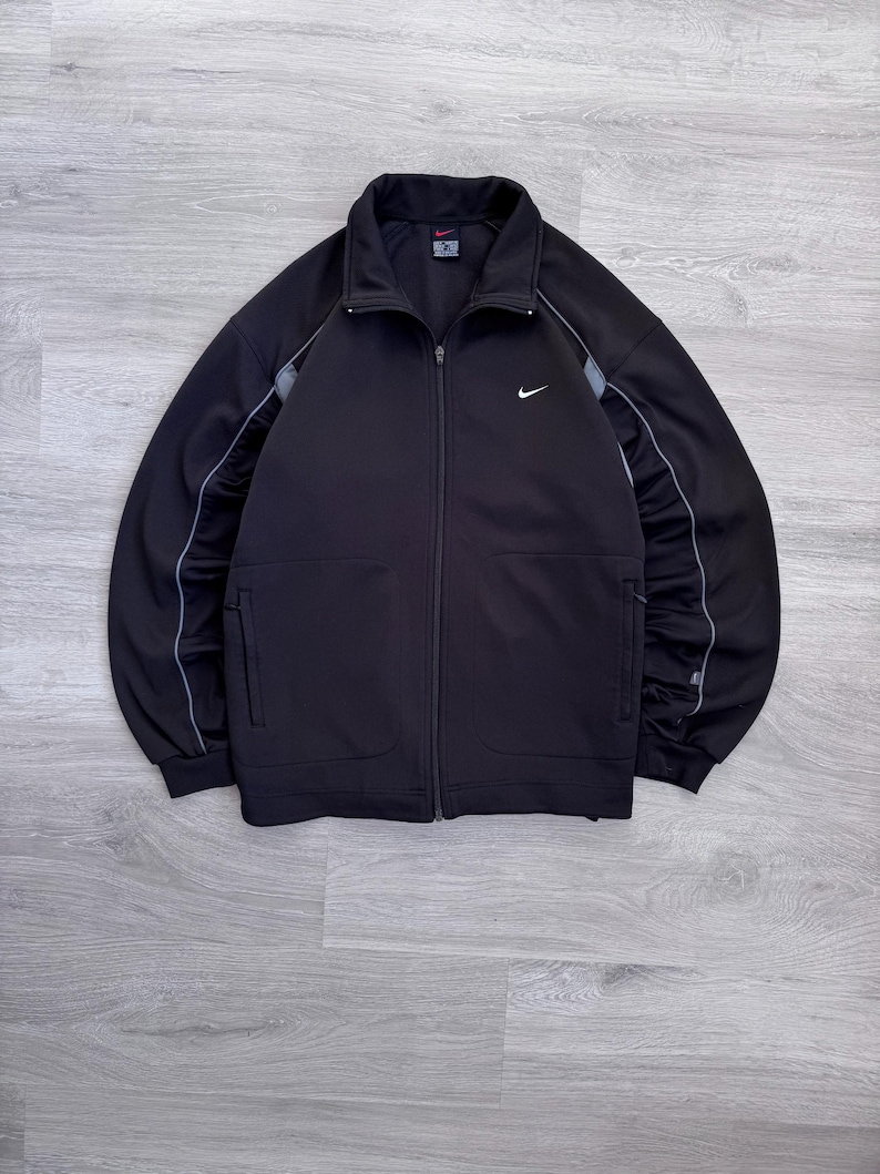 Puede incluir: Chaqueta deportiva negra con cremallera y logotipo blanco de Nike en el pecho. La chaqueta tiene cuello alto, dos bolsillos delanteros y rayas grises en las mangas. Fabricada con un material suave y deportivo.