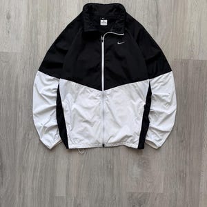 Peut inclure: Veste coupe-vent noire et blanche avec fermeture &eacute;clair compl&egrave;te. La veste pr&eacute;sente une section sup&eacute;rieure noire et une section inf&eacute;rieure blanche, avec un petit logo Nike sur la poitrine. Elle a un col montant et est faite d'un mat&eacute;riau l&eacute;ger.