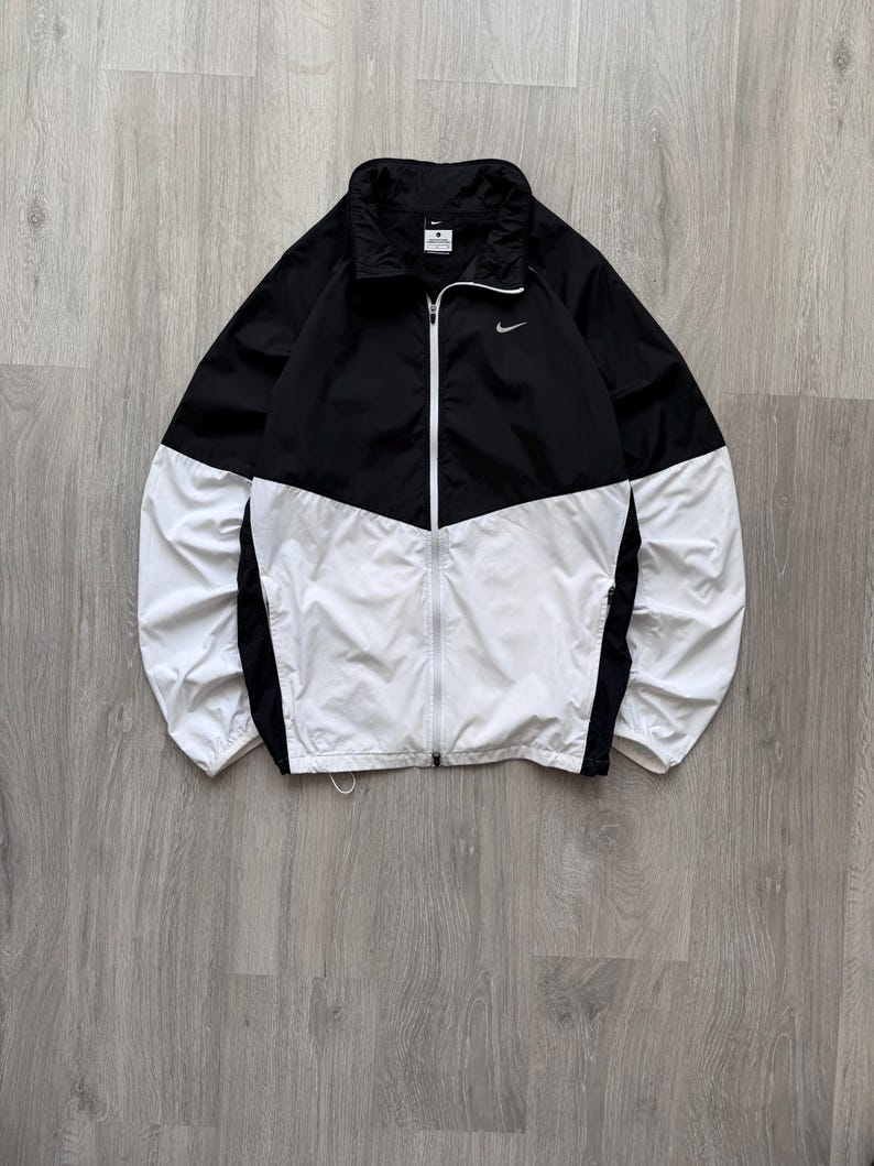 Peut inclure: Veste coupe-vent noire et blanche avec fermeture &eacute;clair compl&egrave;te. La veste pr&eacute;sente une section sup&eacute;rieure noire et une section inf&eacute;rieure blanche, avec un petit logo Nike sur la poitrine. Elle a un col montant et est faite d'un mat&eacute;riau l&eacute;ger.