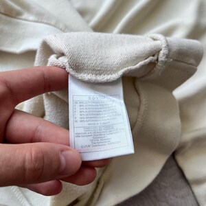 Puede incluir: Primer plano de una etiqueta de sudadera crema sostenida por una mano. La etiqueta muestra texto en varios idiomas, incluidos ingl&eacute;s, espa&ntilde;ol y franc&eacute;s, que detalla la composici&oacute;n de la tela. La tela de la sudadera es visible en el fondo.