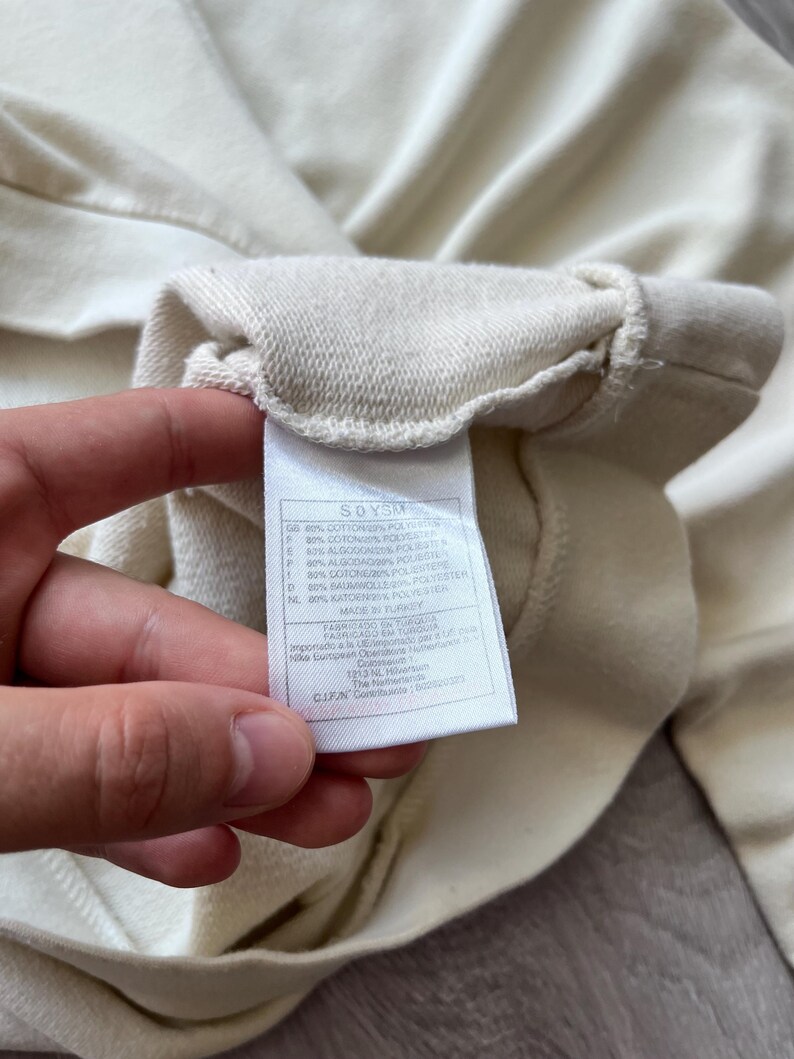 Puede incluir: Primer plano de una etiqueta de sudadera crema sostenida por una mano. La etiqueta muestra texto en varios idiomas, incluidos ingl&eacute;s, espa&ntilde;ol y franc&eacute;s, que detalla la composici&oacute;n de la tela. La tela de la sudadera es visible en el fondo.