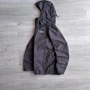Könnte beinhalten: Dunkelgraue Kapuzenjacke mit Reißverschluss und Markenlogo auf der Brust. Die Jacke hat eine Kapuze mit blauen Kordelzügen und eine kleine Tasche. Hergestellt aus leichtem Material, ideal für Outdoor-Aktivitäten.