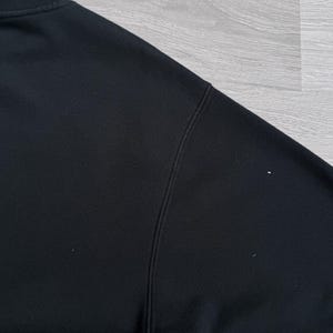 Peut inclure: Gros plan d'un sweat-shirt noir, montrant l'&eacute;paule et l'encolure. Le tissu semble &ecirc;tre une mati&egrave;re douce. Les coutures sont visibles le long des coutures. L'arri&egrave;re-plan est une surface en bois gris clair.