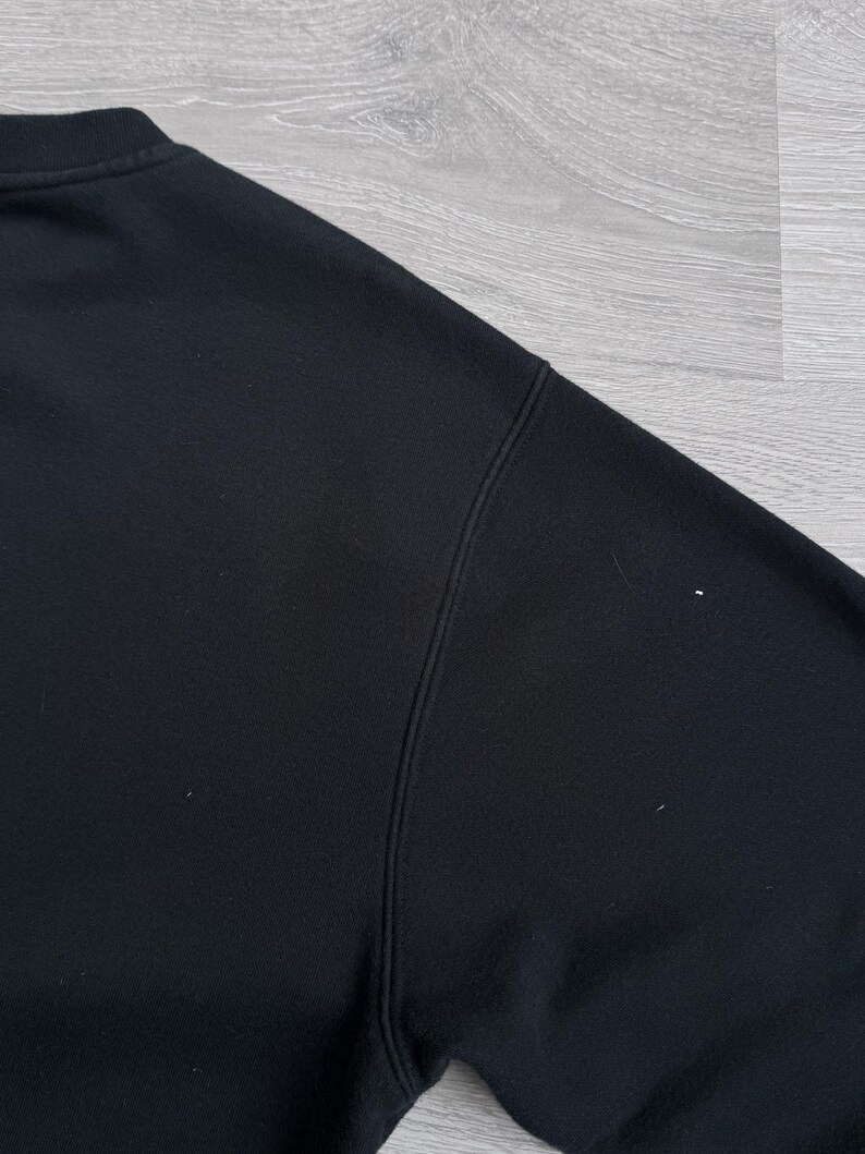 Peut inclure: Gros plan d'un sweat-shirt noir, montrant l'&eacute;paule et l'encolure. Le tissu semble &ecirc;tre une mati&egrave;re douce. Les coutures sont visibles le long des coutures. L'arri&egrave;re-plan est une surface en bois gris clair.