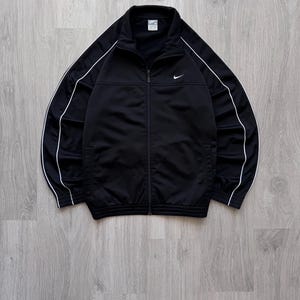 May include: Black track jacket with white stripes down the sleeves and a small Nike logo on the chest. The jacket has a full zipper and a stand-up collar. A classic athletic apparel piece.