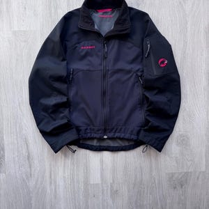 May include: A black and navy blue jacket with a full-length zipper, featuring the brand name "MAMMUT" in pink lettering. The jacket has a high collar, multiple pockets, and a pink logo on the sleeve. The jacket is designed for outdoor activities.