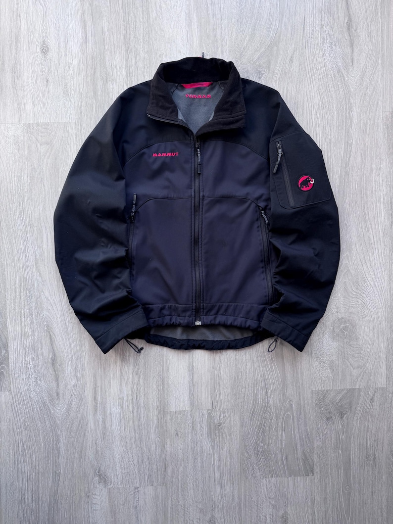 May include: A black and navy blue jacket with a full-length zipper, featuring the brand name "MAMMUT" in pink lettering. The jacket has a high collar, multiple pockets, and a pink logo on the sleeve. The jacket is designed for outdoor activities.