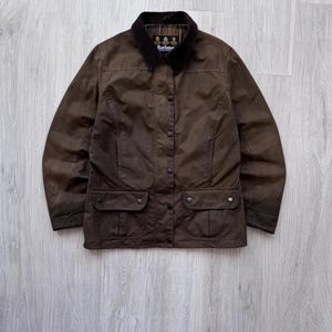 Puede incluir: Chaqueta marrón de algodón encerado con cuello de pana y cuatro bolsillos delanteros. La chaqueta tiene cierre de botones y un diseño clásico. El logo de Barbour es visible en el cuello.