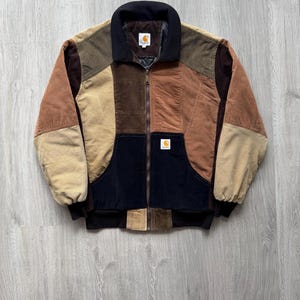 May include: A multi-colored corduroy jacket with a black collar and zipper. The jacket features a patchwork design with various shades of brown, tan, and black. The Carhartt logo is visible on the chest pocket and inside the collar.
