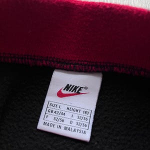 May include: A black garment with a red collar, close-up view. A white label features the Nike logo and size details: Size L, Height 183cm, GB 42/44, F 52/56, I 52/56, D 52/56. The label reads "Made in Malaysia."