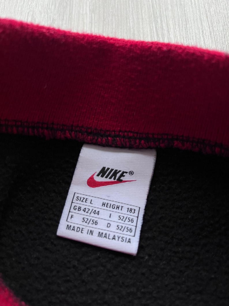 May include: A black garment with a red collar, close-up view. A white label features the Nike logo and size details: Size L, Height 183cm, GB 42/44, F 52/56, I 52/56, D 52/56. The label reads "Made in Malaysia."