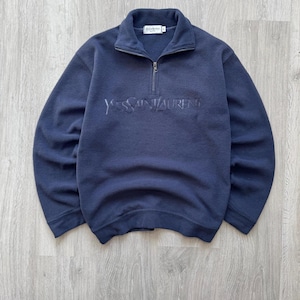 Op de afbeelding: Marineblau fleece trui met een kwart rits sluiting en de in reliëf aangebrachte tekst "Yves Saint Laurent" over de borst. De kraag is een opstaande kraag stijl. Het kledingstuk is een medium maat.