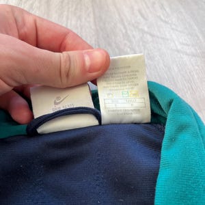 May include: A close-up of a navy blue and teal athletic jacket. The jacket has a white tag with the Nike logo and text. The tag also includes washing instructions and size information. The jacket is made of polyester.