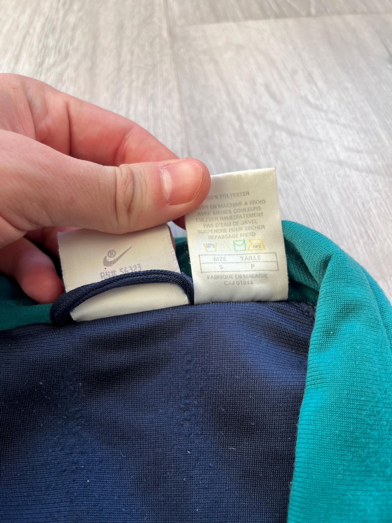 May include: A close-up of a navy blue and teal athletic jacket. The jacket has a white tag with the Nike logo and text. The tag also includes washing instructions and size information. The jacket is made of polyester.