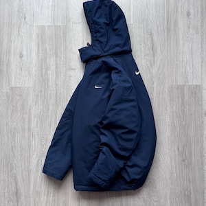 May include: A navy blue hooded jacket with a white Nike logo on the chest and back. The jacket features a full zip and a drawstring hood. The jacket is made of a lightweight material and appears to be water-resistant.