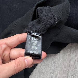 Peut inclure: Sweat-shirt FILA noir avec lettrage blanc. L'image montre un gros plan de l'&eacute;tiquette du v&ecirc;tement, qui indique la composition du tissu comme &eacute;tant 80% coton et 20% polyester. L'&eacute;tiquette indique &eacute;galement que le v&ecirc;tement a &eacute;t&eacute; fabriqu&eacute; en Indon&eacute;sie.