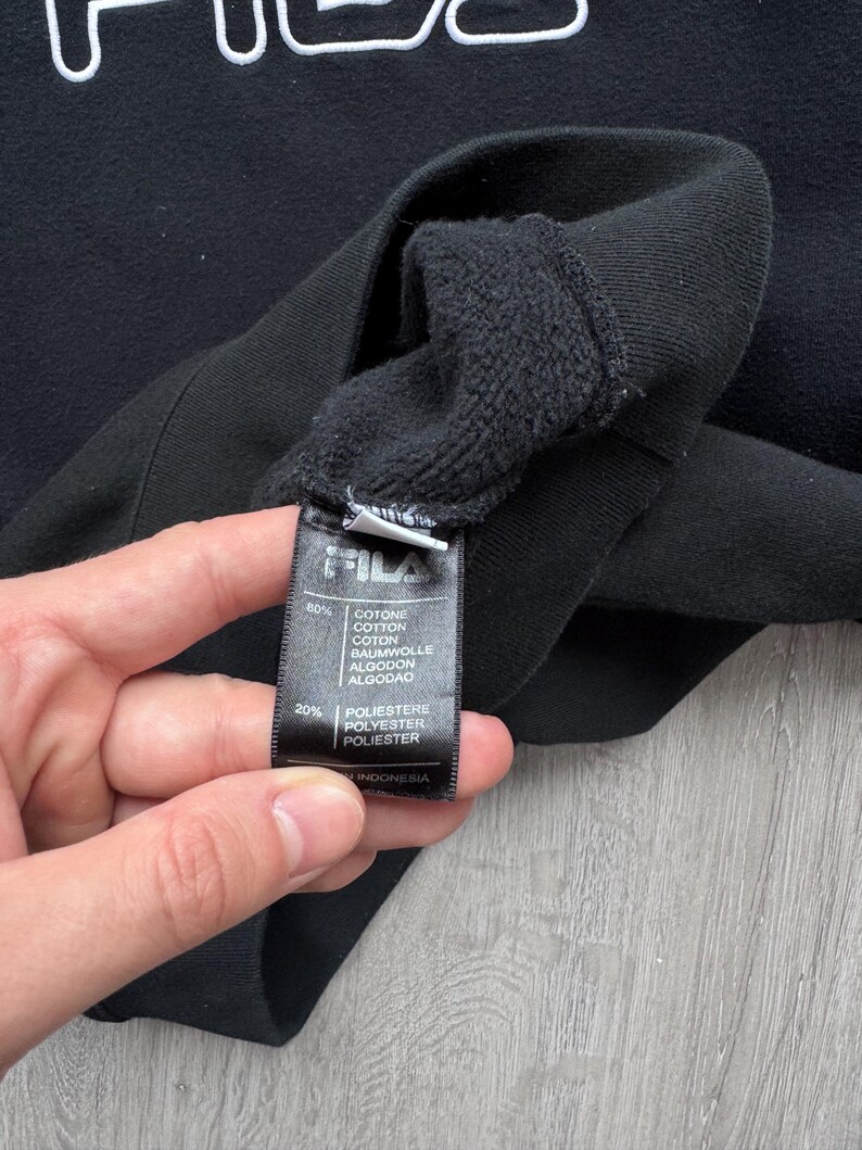 Peut inclure: Sweat-shirt FILA noir avec lettrage blanc. L'image montre un gros plan de l'&eacute;tiquette du v&ecirc;tement, qui indique la composition du tissu comme &eacute;tant 80% coton et 20% polyester. L'&eacute;tiquette indique &eacute;galement que le v&ecirc;tement a &eacute;t&eacute; fabriqu&eacute; en Indon&eacute;sie.