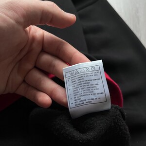 May include: A black garment with a red interior is held up, revealing a tag with washing instructions in multiple languages. The tag includes symbols for washing, drying, and ironing. The garment appears to be made of a soft, dark material.
