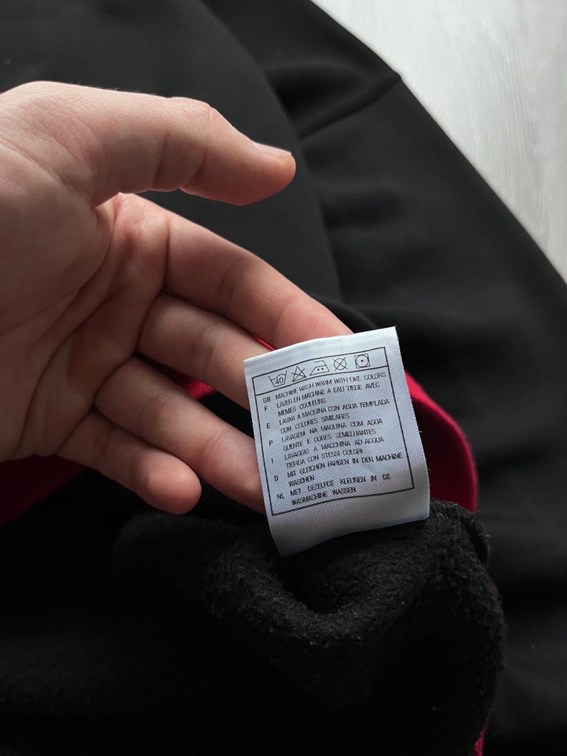May include: A black garment with a red interior is held up, revealing a tag with washing instructions in multiple languages. The tag includes symbols for washing, drying, and ironing. The garment appears to be made of a soft, dark material.
