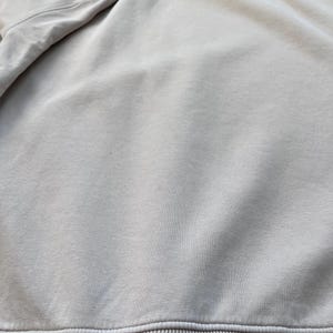 May include: A close-up view of a light beige sweatshirt. The sweatshirt has a ribbed hem and cuffs. The fabric appears to be soft and slightly textured. The sleeves are visible at the top left.