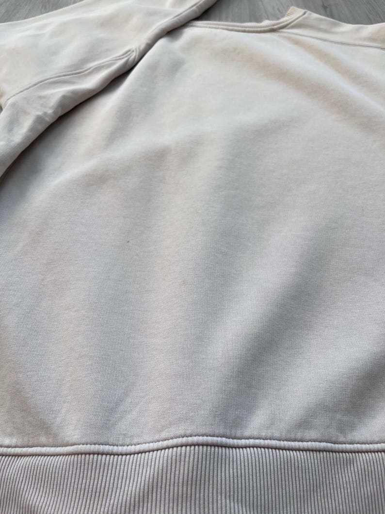 May include: A close-up view of a light beige sweatshirt. The sweatshirt has a ribbed hem and cuffs. The fabric appears to be soft and slightly textured. The sleeves are visible at the top left.