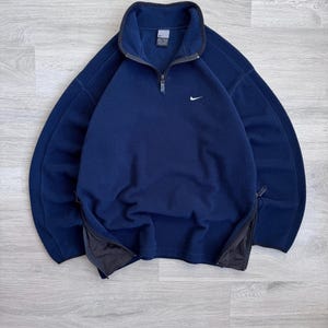 May include: Navy blue fleece pullover with a quarter-zip closure and a small white Nike logo on the chest. Features side zippers and a high collar. A warm and comfortable athletic top.