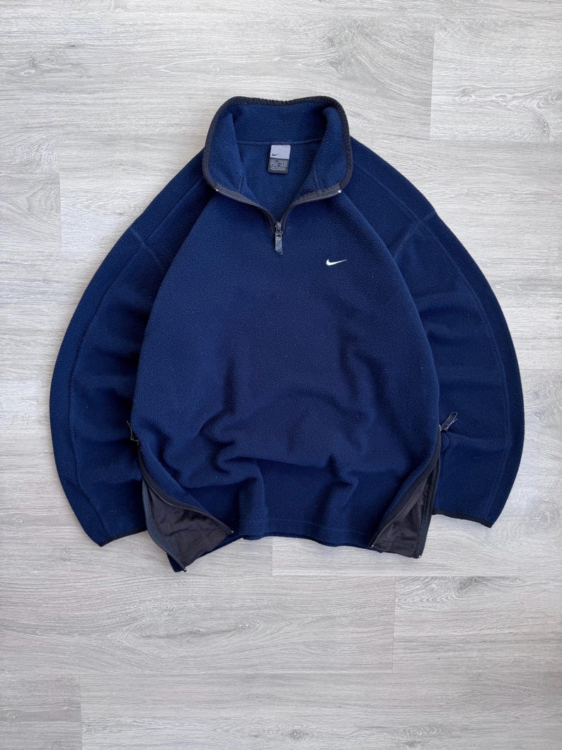 May include: Navy blue fleece pullover with a quarter-zip closure and a small white Nike logo on the chest. Features side zippers and a high collar. A warm and comfortable athletic top.