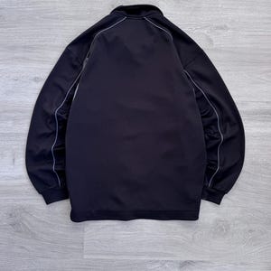 Puede incluir: Chaqueta deportiva negra con cremallera completa y cuello alto. La chaqueta tiene ribetes blancos a lo largo de las costuras y alrededor del cuello. Fabricada con un material liso, posiblemente sint&eacute;tico.