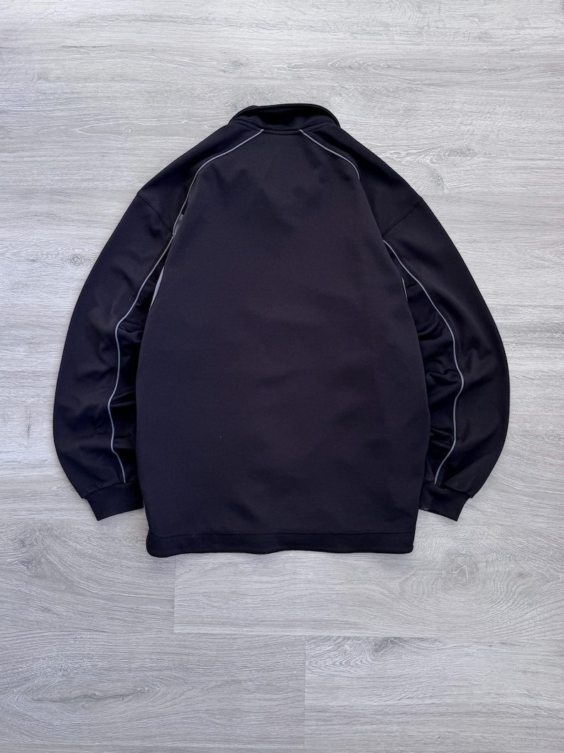 Puede incluir: Chaqueta deportiva negra con cremallera completa y cuello alto. La chaqueta tiene ribetes blancos a lo largo de las costuras y alrededor del cuello. Fabricada con un material liso, posiblemente sint&eacute;tico.