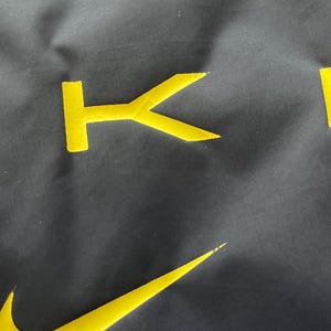 May include: Close-up of a dark gray fabric with yellow embroidered details. The Nike swoosh logo and the letter "K" are visible. The fabric has a slightly textured appearance.