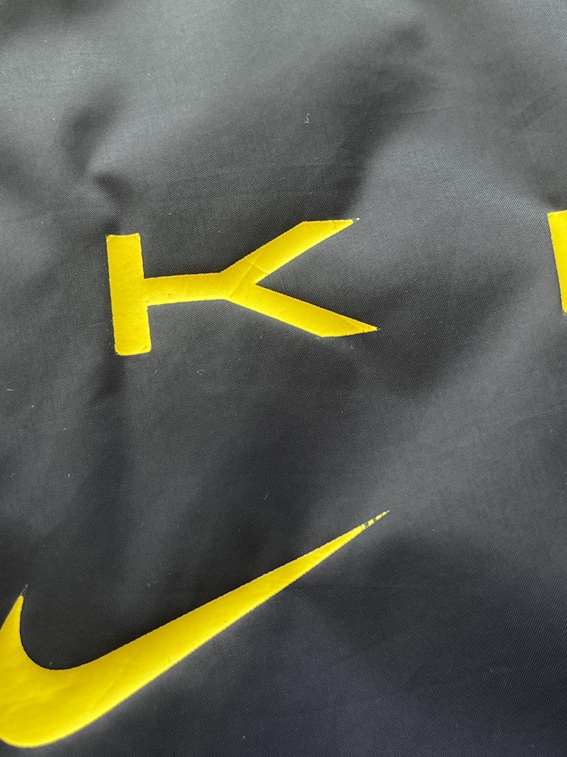 May include: Close-up of a dark gray fabric with yellow embroidered details. The Nike swoosh logo and the letter "K" are visible. The fabric has a slightly textured appearance.
