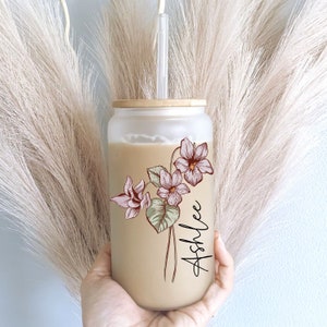 May include: A frosted glass tumbler with a bamboo lid and a straw. The tumbler has a floral design with pink flowers and green leaves. The name "Ashlee" is written in cursive on the side of the tumbler.