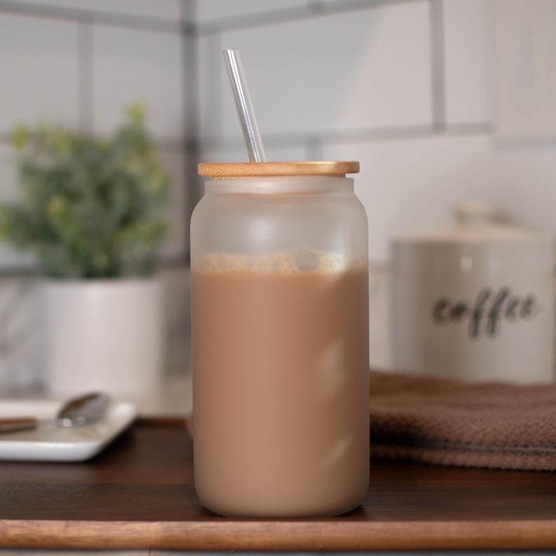 May include: A frosted glass jar with a brown lid and a clear glass straw. The jar is filled with a brown liquid and ice cubes.
