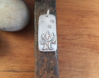 Puede incluir: Un colgante de plata con un diseño de árbol y cinco estrellas. El colgante es rectangular y tiene una superficie texturizada.