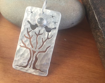 Puede incluir: Colgante rectangular de plata con diseño de árbol y pájaros. El colgante presenta una textura martillada, una silueta de árbol, dos recortes de pájaros y recortes en forma de estrella. Una pequeña piedra redonda está engastada cerca de la parte superior.