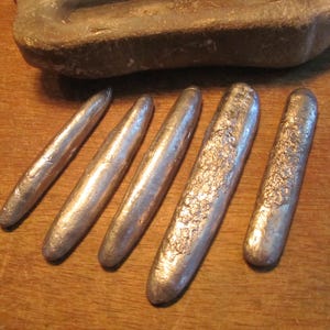 May include: Five elongated, silver-coloured metal ingots with rounded ends arranged in a row. The surfaces of the ingots vary in texture, with some appearing smooth and others showing a rough, crystalline, or pitted finish. A grey stone mould sits above.