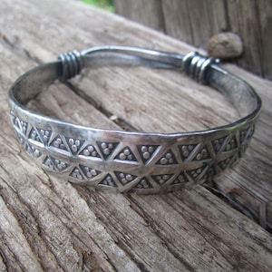 May include: Silver-toned bangle bracelet with a geometric triangle and dot pattern. The bracelet has a hinged design and is displayed on a weathered wooden surface. The bracelet's design is repeated around the band.