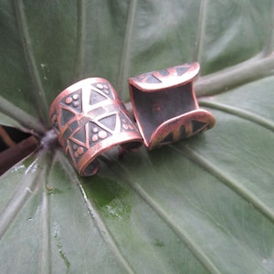 May include: Two copper-colored cuff rings with geometric designs. The rings have a dark patina in the recessed areas, and are set against a large, green leaf. The rings are open at the back.