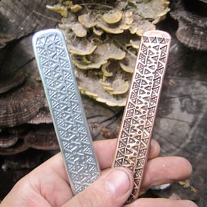Puede incluir: Dos marcapáginas rectangulares de metal, uno en tono plateado y otro en tono cobre, ambos con un patrón geométrico idéntico en relieve de triángulos y puntos dispuestos en columnas verticales por toda la superficie de cada pieza.