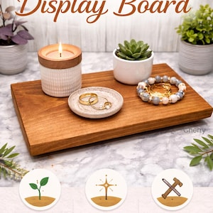 May include: A rectangular cherry wood display board featuring a lit candle in a white textured container, a small speckled ceramic dish holding gold rings, a succulent in a white pot, and a beaded bracelet. Text reads HANDMADE SOLID WOOD Display Board.